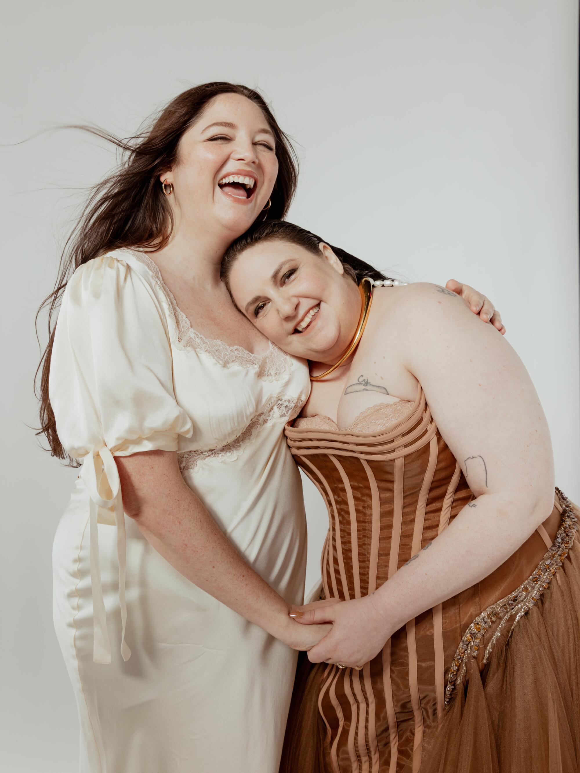 A smiling woman in a white dress hugs another woman in a brown corset dress.