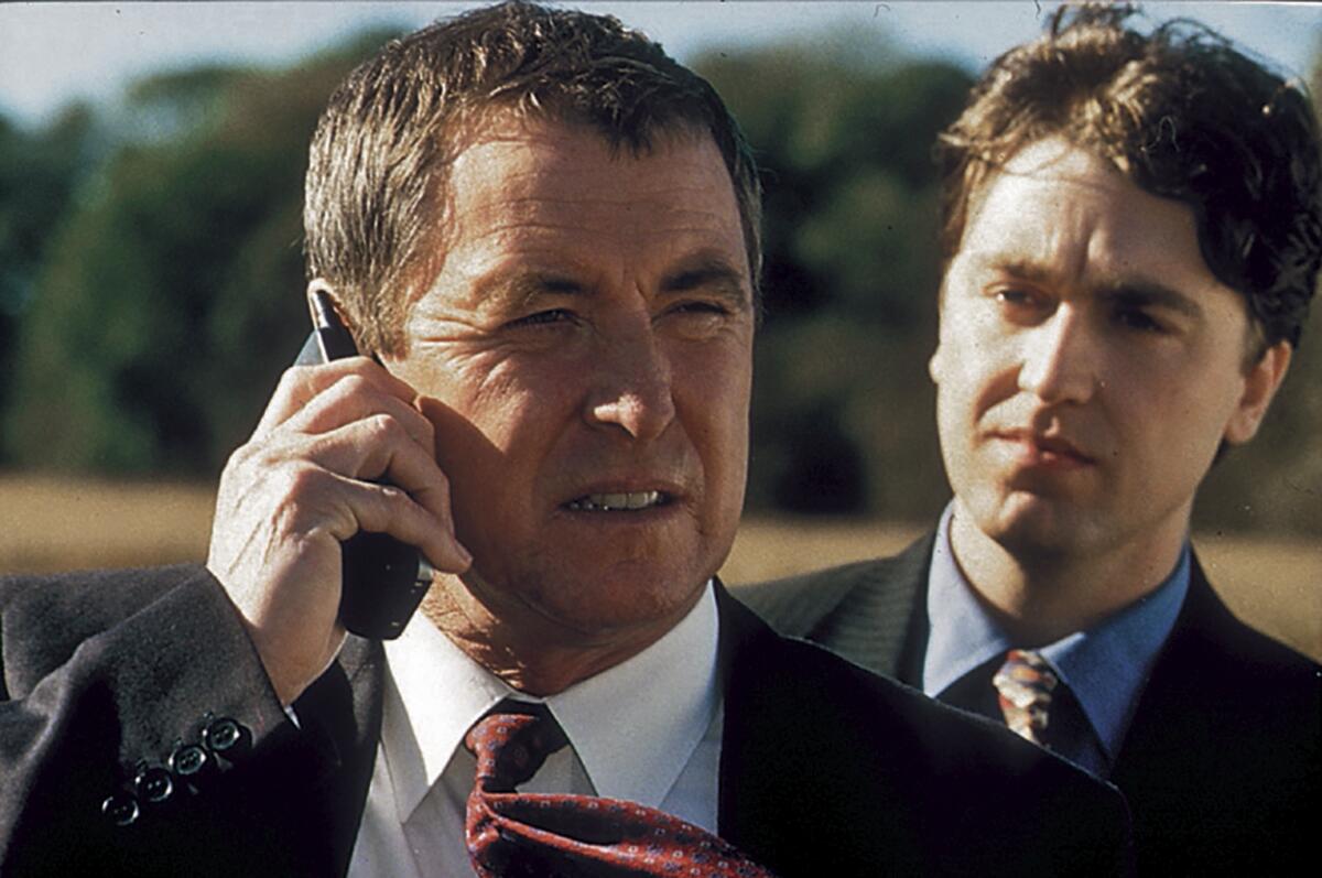 A young man stands behind and looks at an older man using a mobile phone.