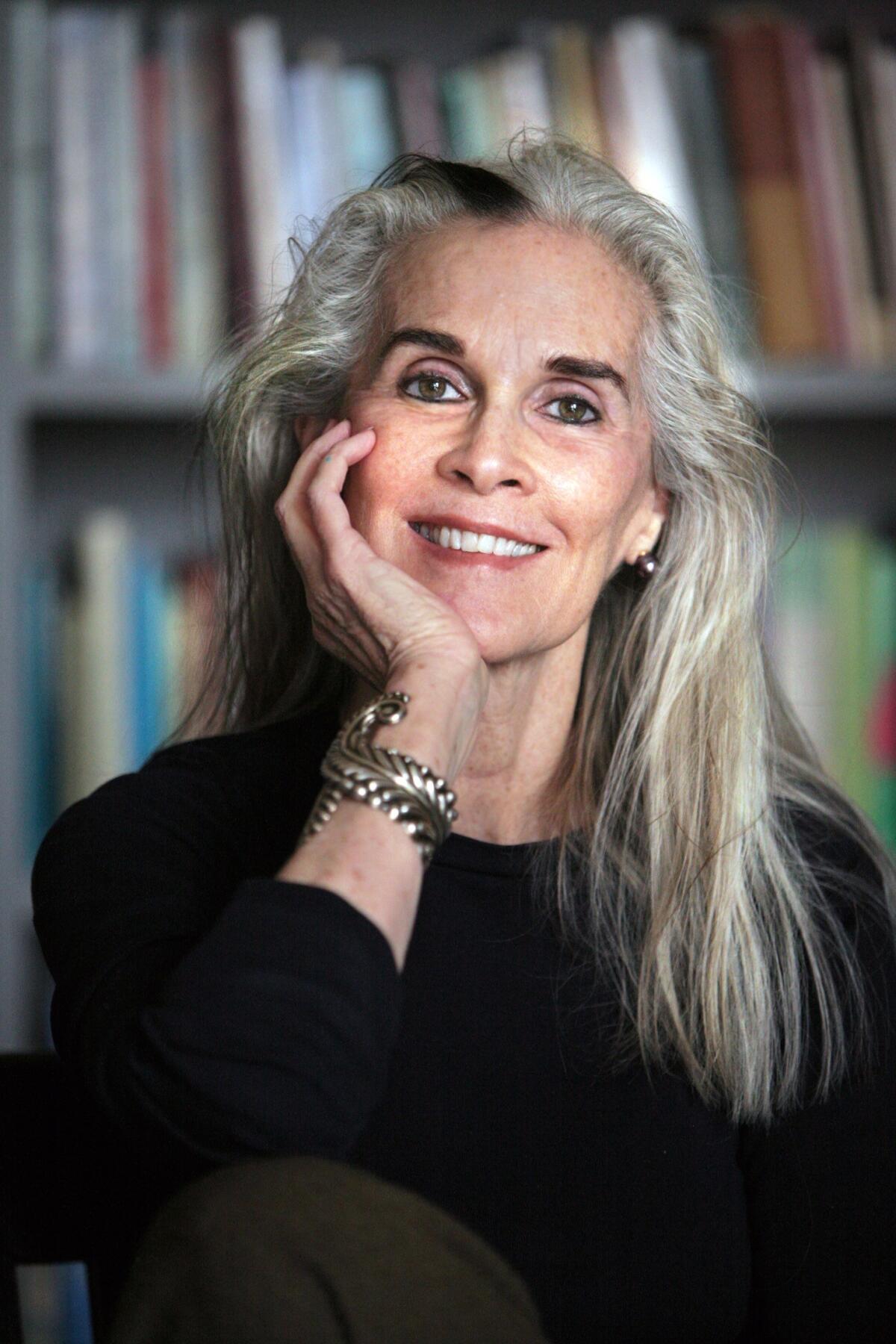 A woman with long gray hair and a long-sleeved black shirt in front of a bookshelf.