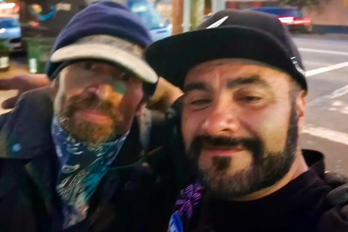 A selfie of two bearded men, both wearing caps and one with a scarf, as they stand in an urban area