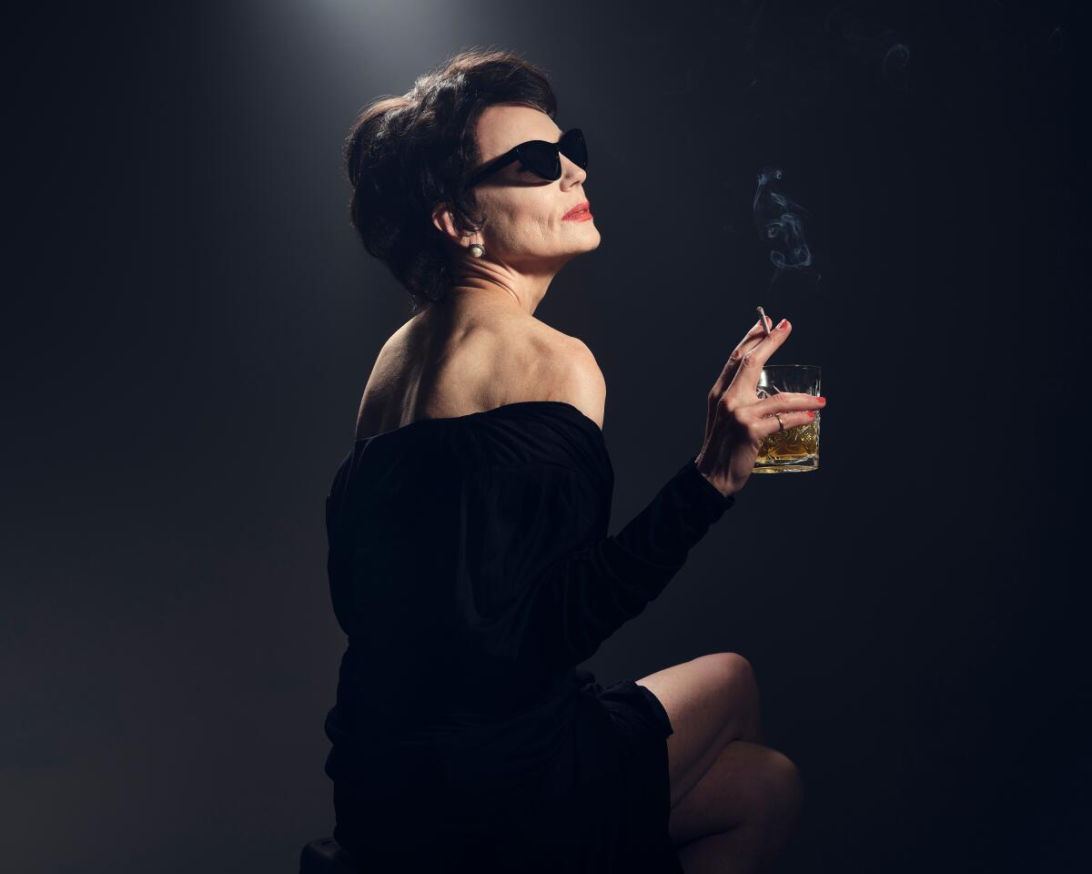 An elegant woman in a black off-the-shoulder dress and sunglasses holds a drink and lit cigarette in the same hand 