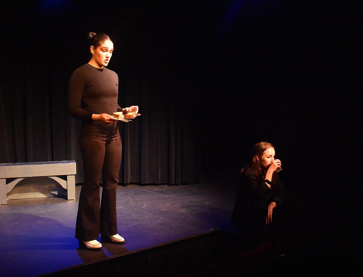 A woman in black stands onstage while another sits at the edge.