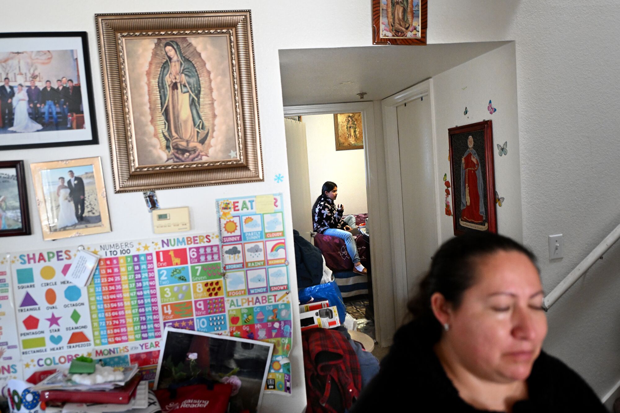 A girl in the background sits in a room as her mother stands in the foreground.