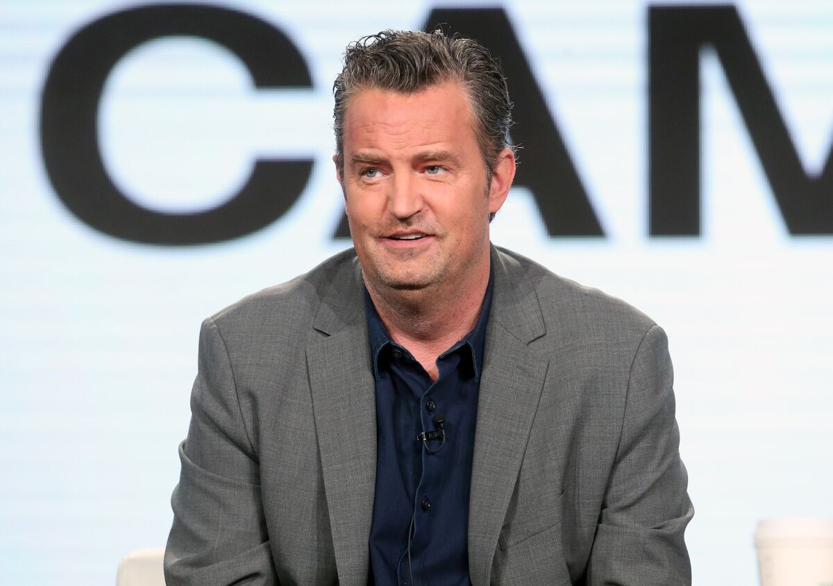 Matthew Perry sitting in a gray suit and dark shirt