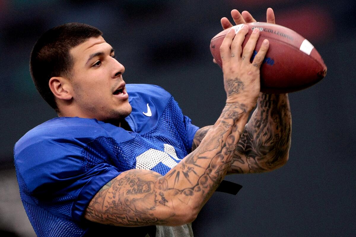 A football player with heavily tattooed arms, wearing a blue shirt and shin guards, holding a football in his hands close to his face.