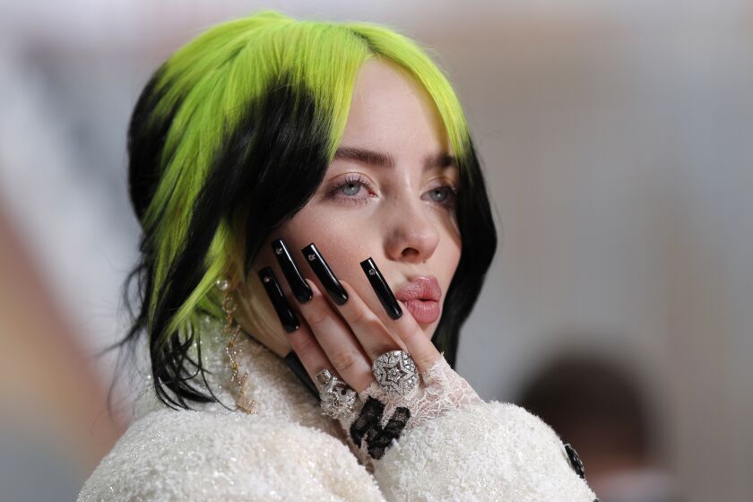 A woman with green and black hair raising a hand to her face with long black nails