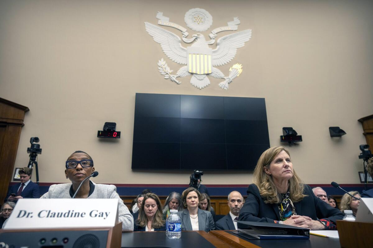 A Black woman and a white woman sit before microphones as others sit behind them listening.