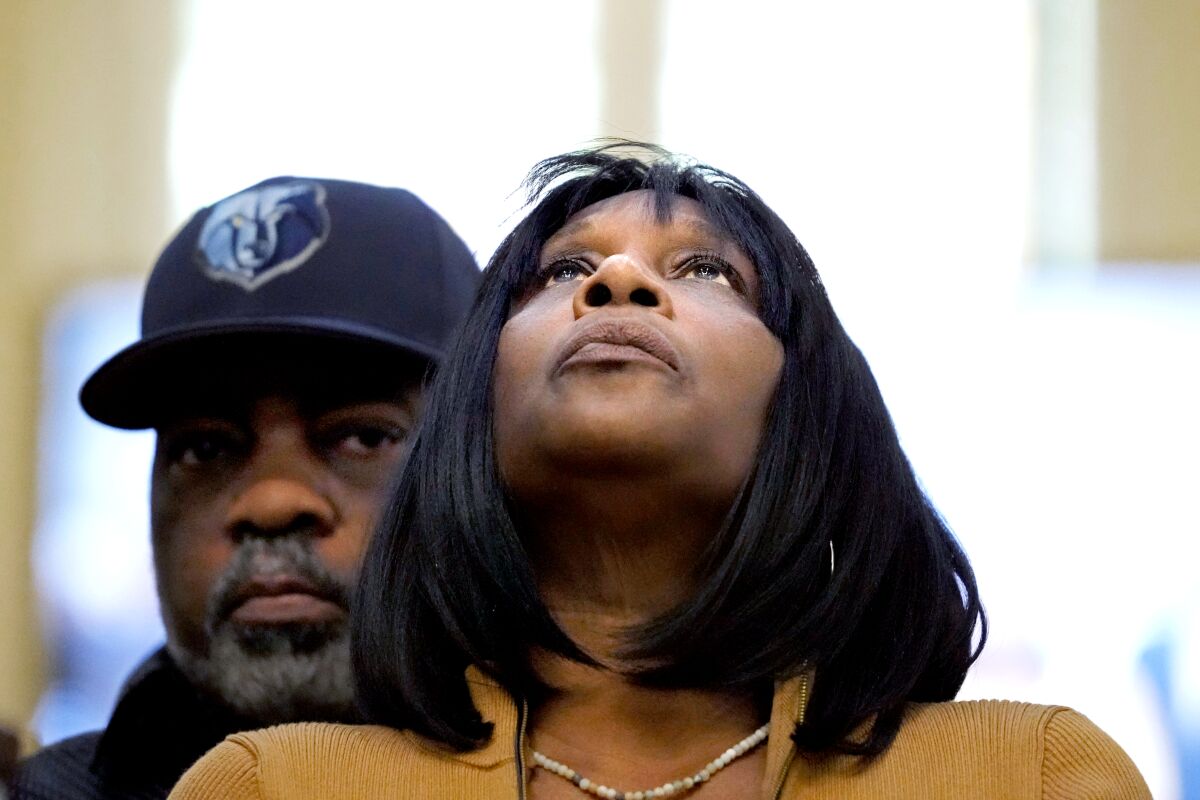 Une femme aux cheveux noirs mi-longs penchant la tête et regardant vers le haut. Un homme coiffé d'une casquette se tient derrière elle.