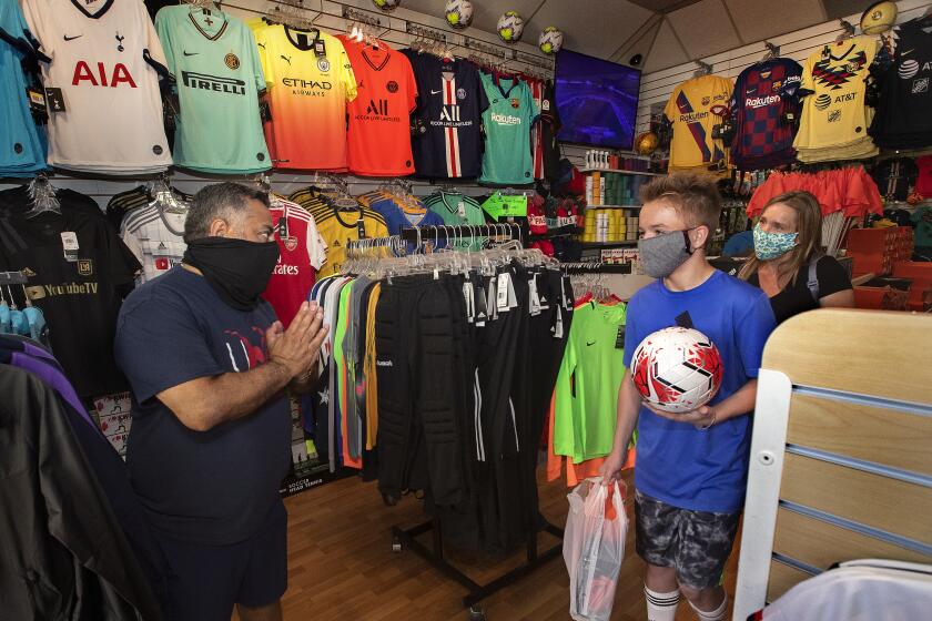 NEWHALL, CA-MAY 13, 2020: Carlos Marroquin, 53, left, owner of Planet Soccer, shows his appreciation to customers Benton Watkins, 13, and his mother Noelle,47, of Santa Clarita, after she purchased a soccer ball, soccer cleats and shin guards for he son at the store in Newhall that re-opened. All are wearing protective covering against the coronavirus. Noelle and Benton have been customers since he was 8 years old. Marroquin has owned the store for the past 16 years and during that time, he has provided gear but more importantly, support and advice to AYSO leagues, high school teams and weekend warriors in the Santa Clarita Valley. (Mel Melcon/Los Angeles Times)