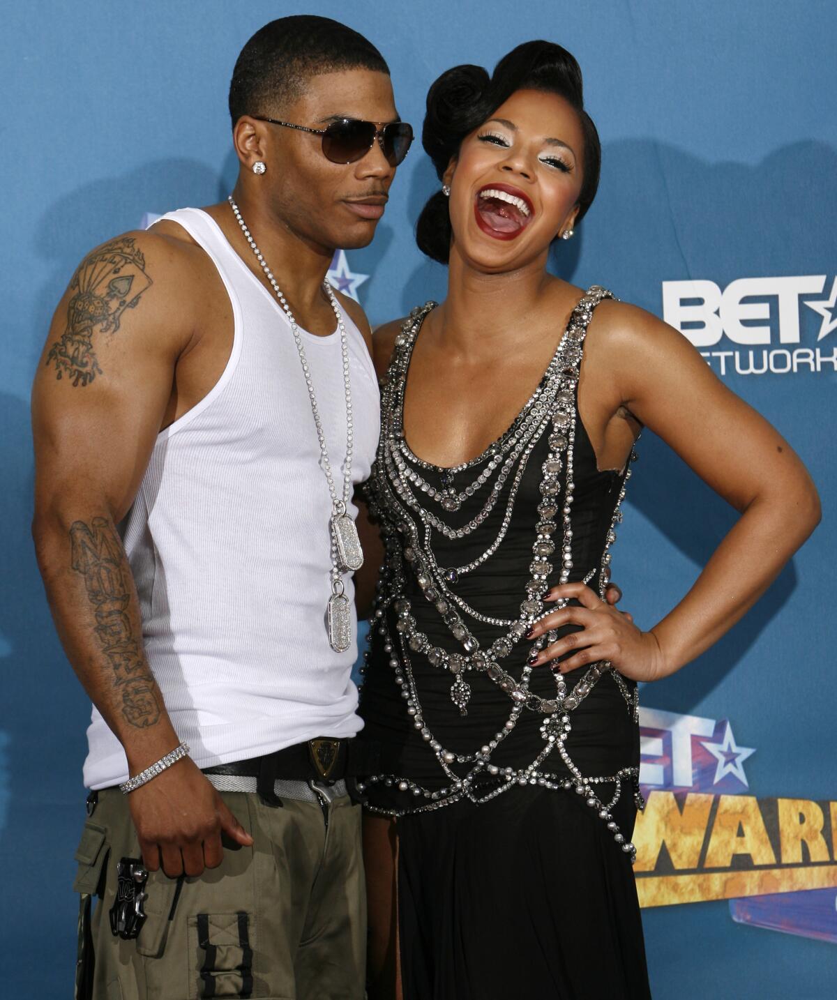 Nelly, left, wears a white A-shirt and olive green pants and Ashanti, right, wears a black dress wrapped in silver jewelry 