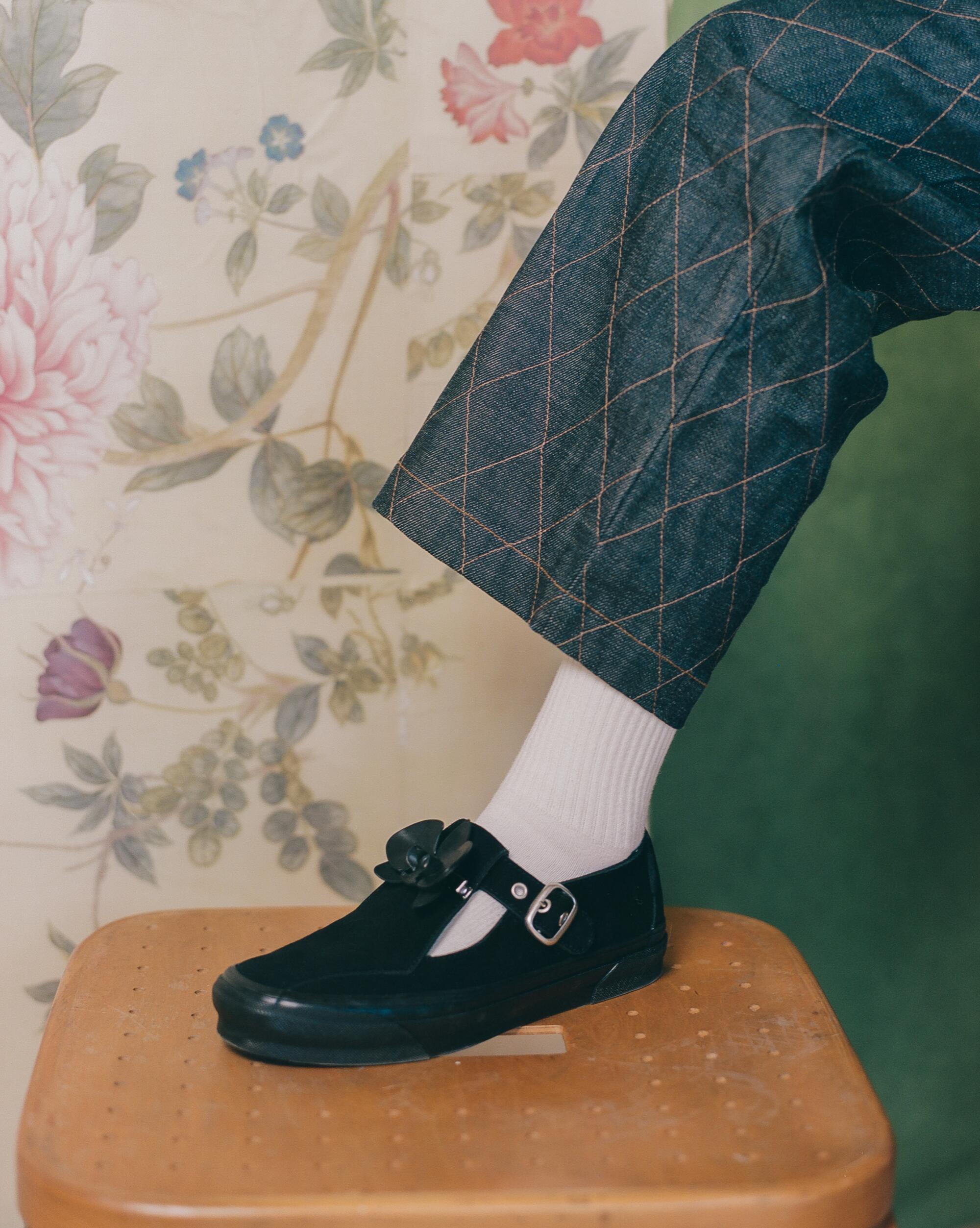 A close-up image of a person's leg propped on a stool, showing off a pair of black Mary Janes with an orchid.