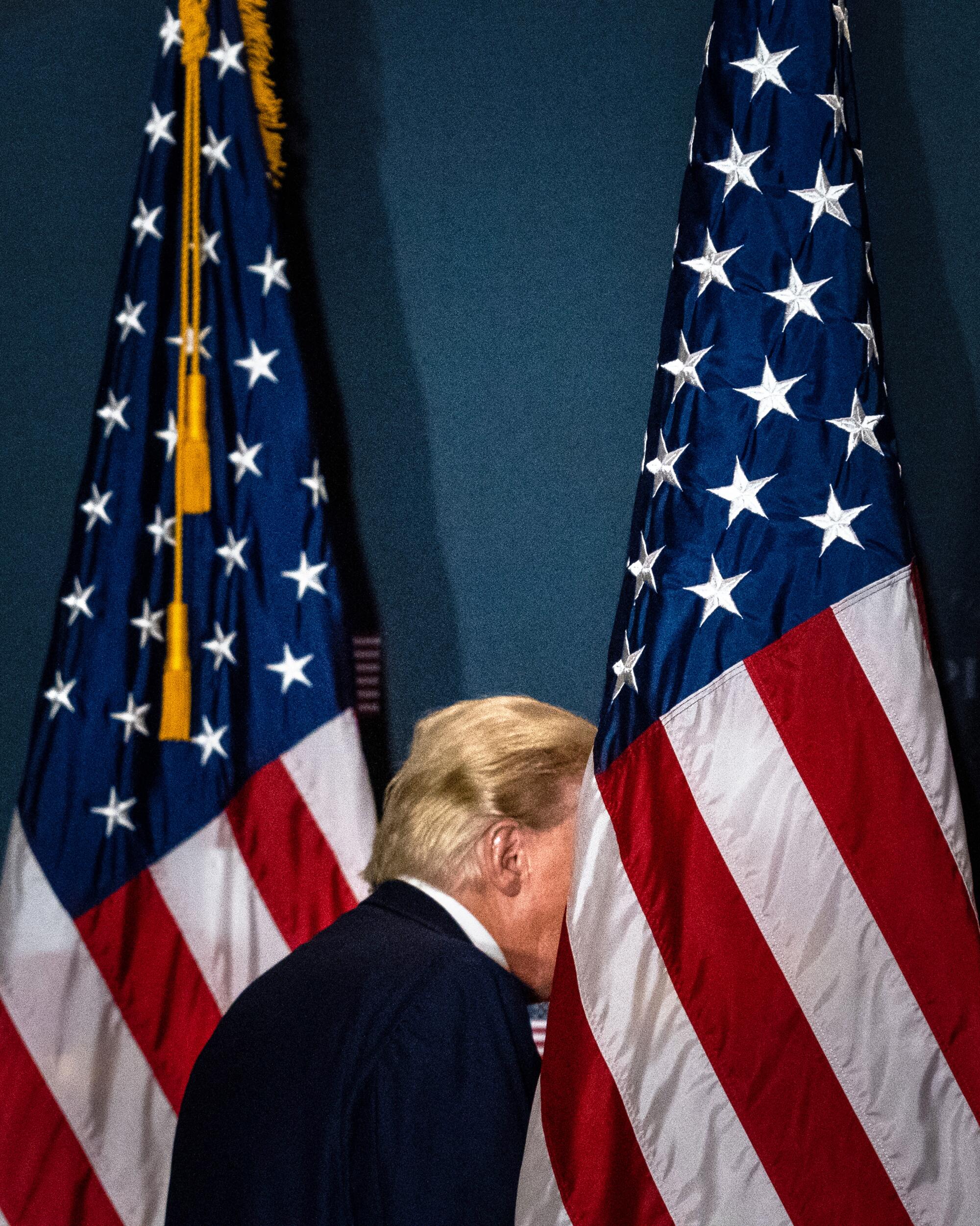 A vertical frame of Donald Trump walking in between two American flags - his face obstructed by the flag at right.