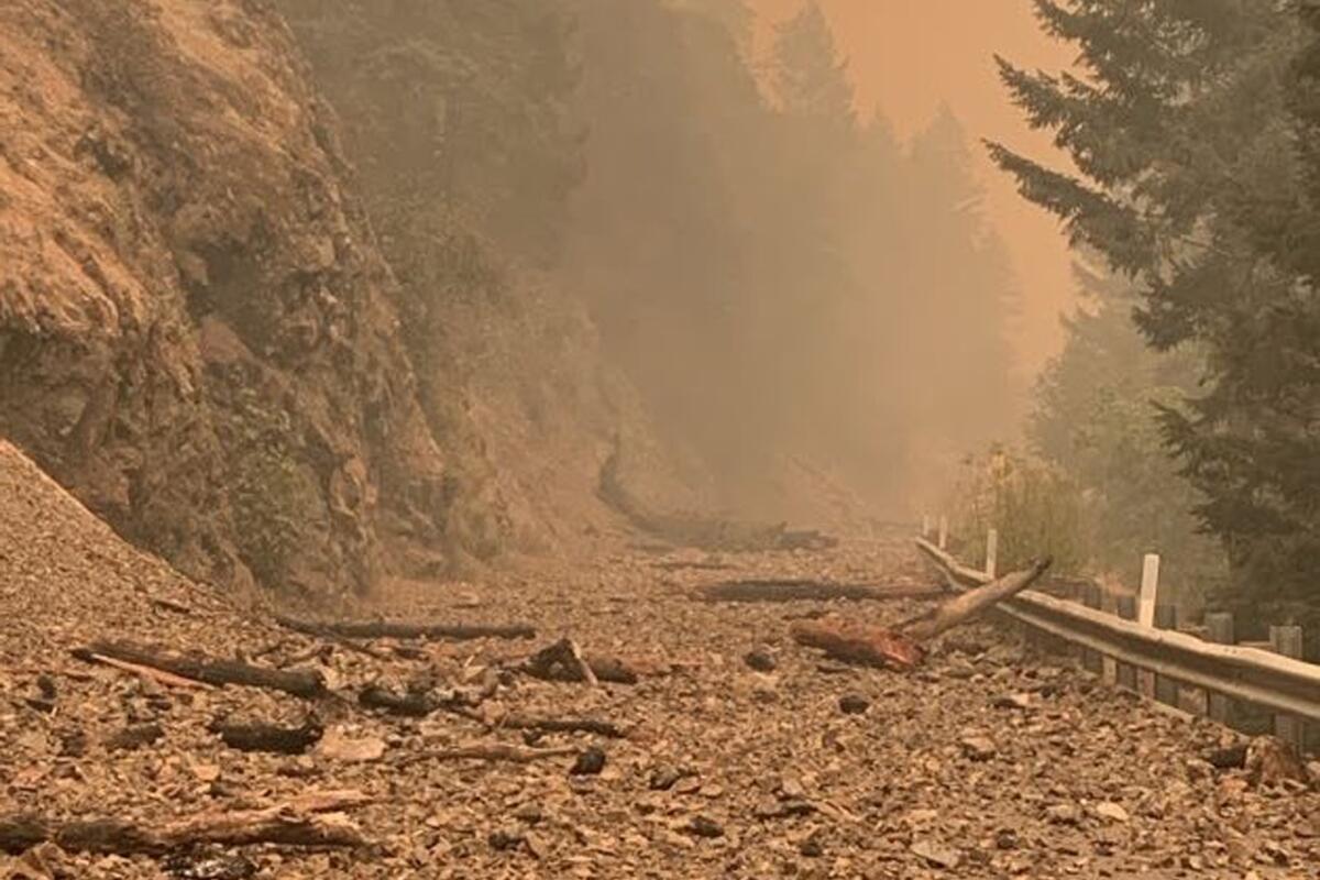 Debris covers a closed road in a mountainous forest area with thick smoke in the sky.