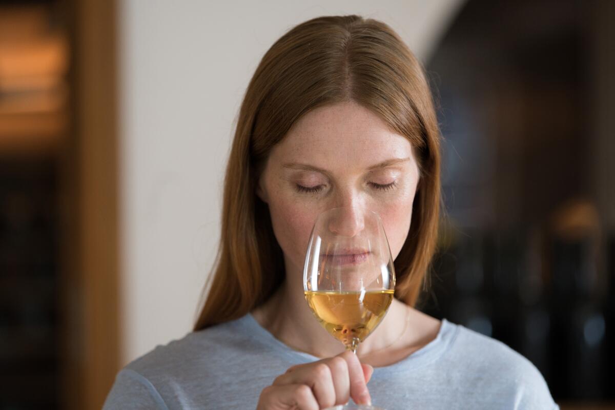 A woman holds a glass of white wine up beneath her nose.