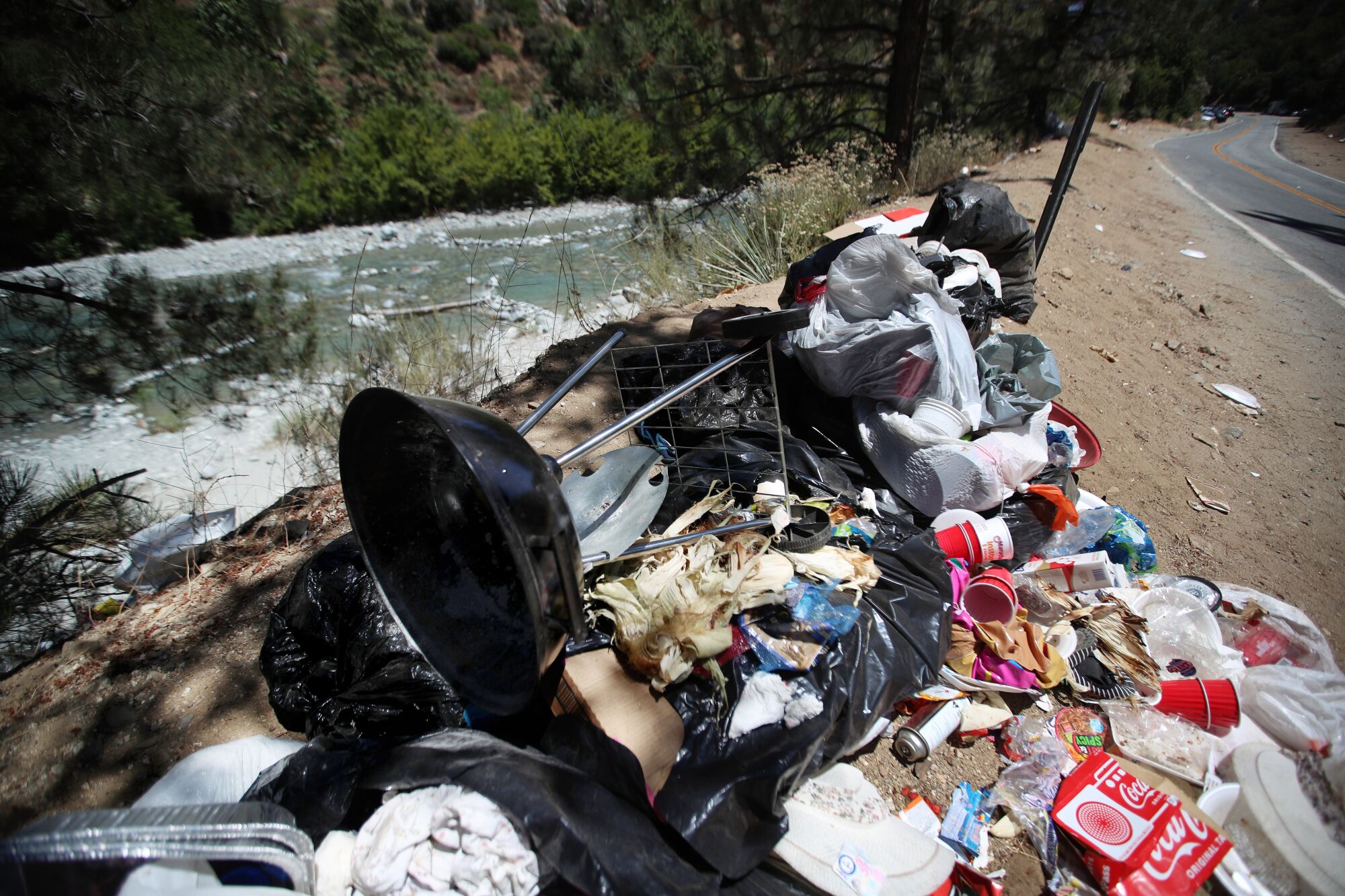 A discarded BBQ grill rests atop a heap of trash beside a river.