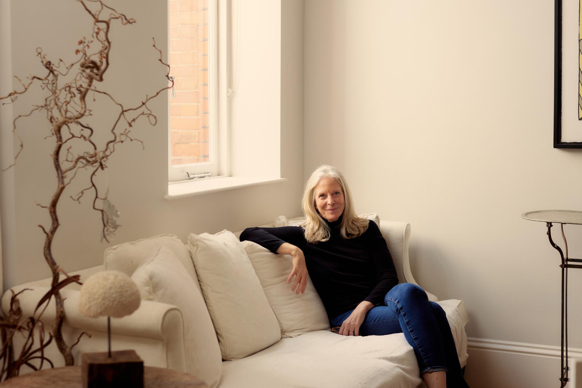 Bonnie Garmus, in a black turtleneck and blue jeans, sits on a white couch in her home.