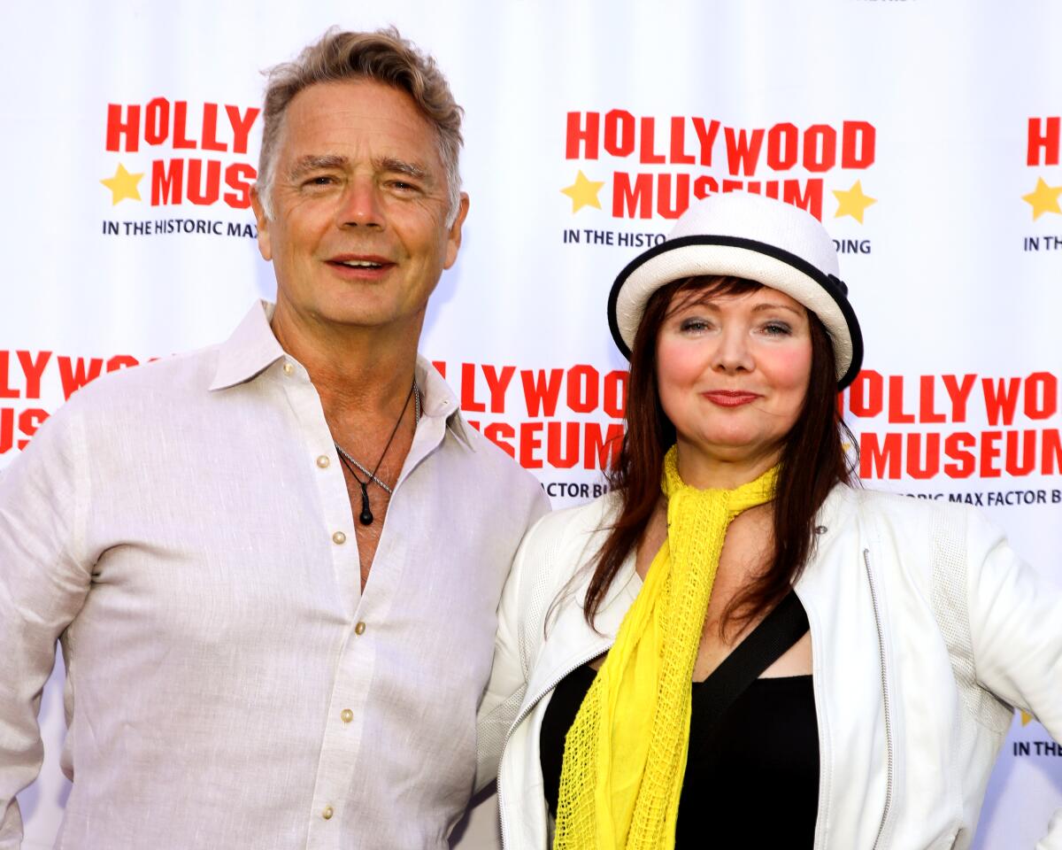 John Schneider posing in a dress shirt with Dee Dee Sorvino wearing a white hat and yellow scarf