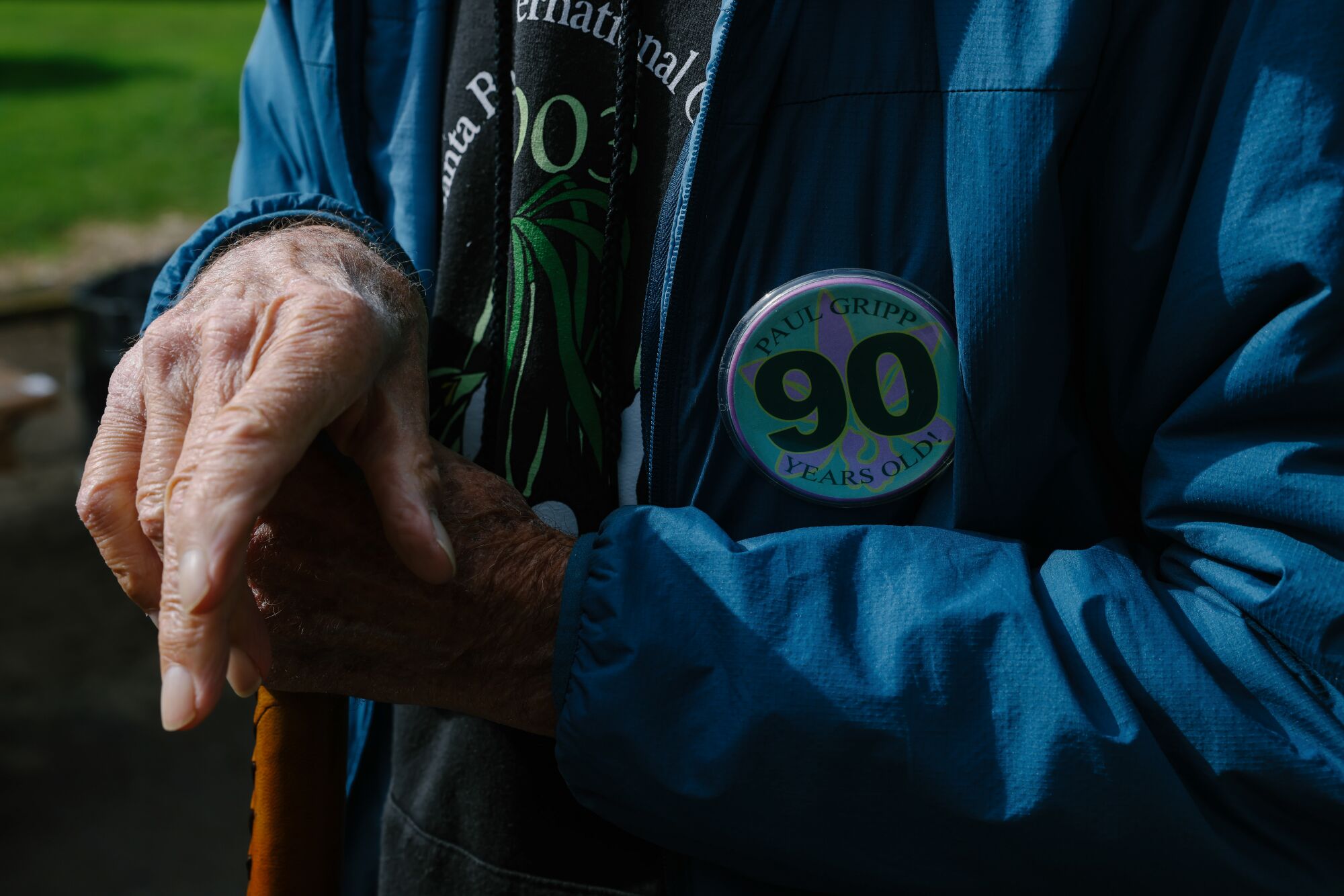 A closeup of a man's hand resting on a cane; his jacket has a button that says 90