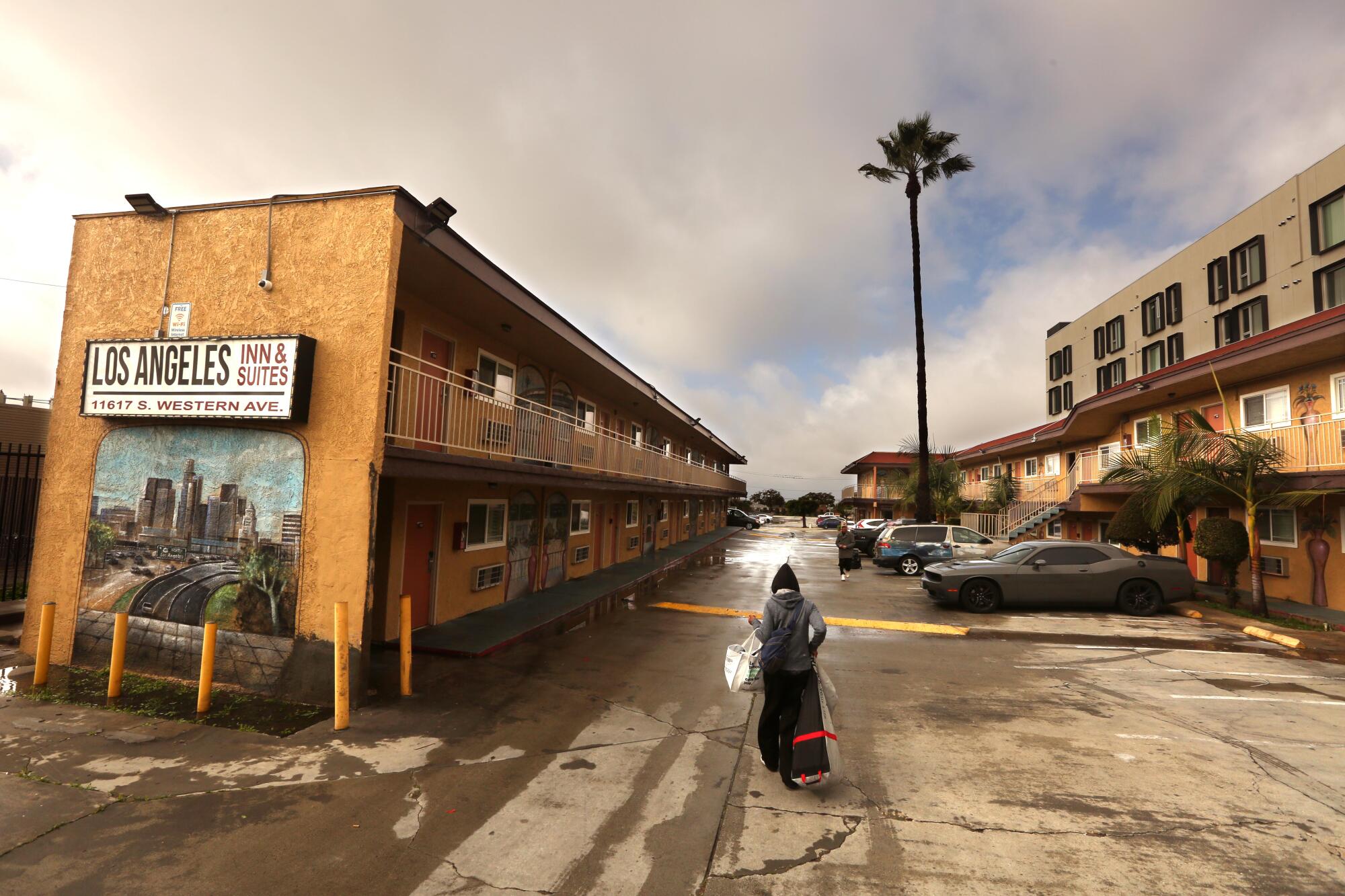 A person, seen from behind, carries bags while walking alongside a building with a sign that says Los Angeles Inn & Suites