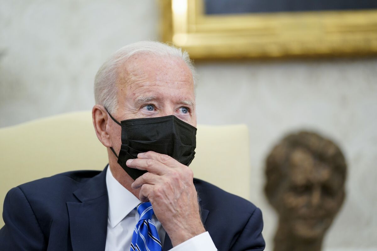 President Biden, wearing a mask, puts his hand to his face while listening.