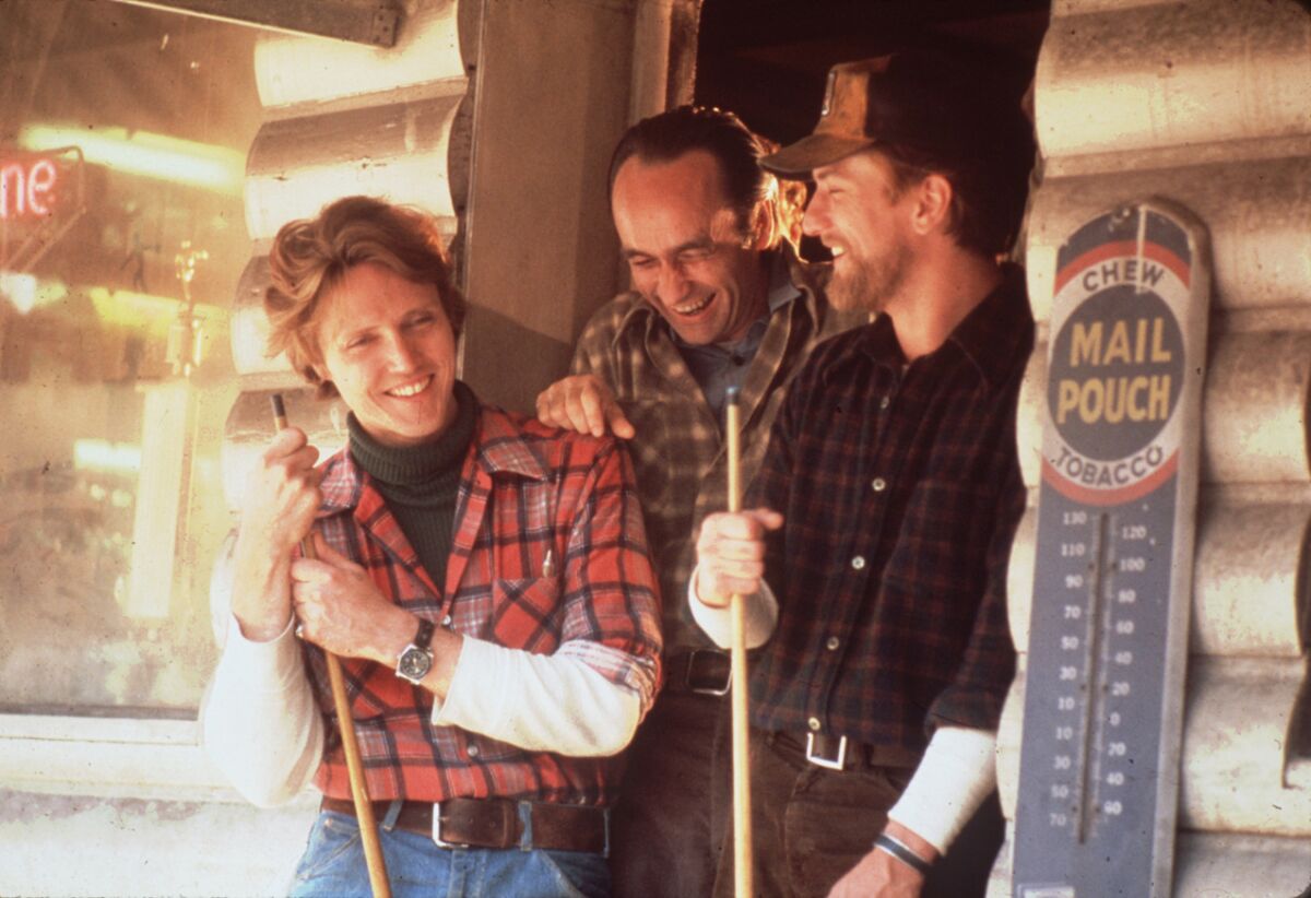 Three men in flannel shirts stand laughing in a doorway, with two of them holding  pool cues.