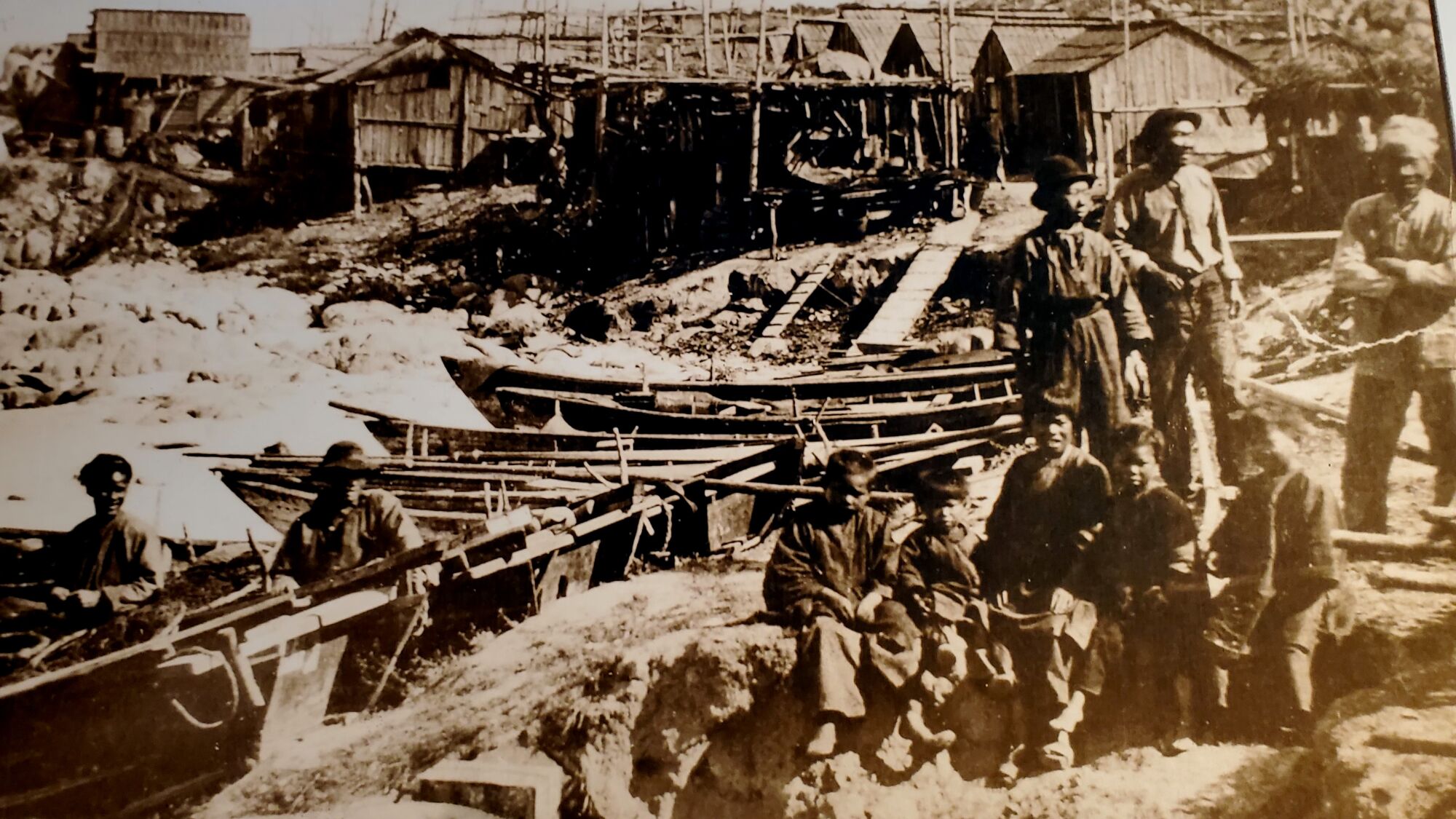 A historic black-and-white photo of fishermen and children of Chinese descent near a cluster of wooden boats and houses.
