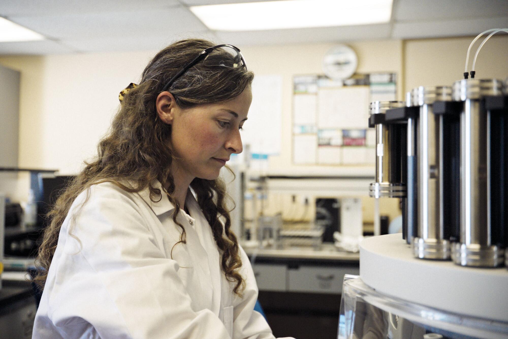 A woman in a white lab coat stands next to some scientific equipment.