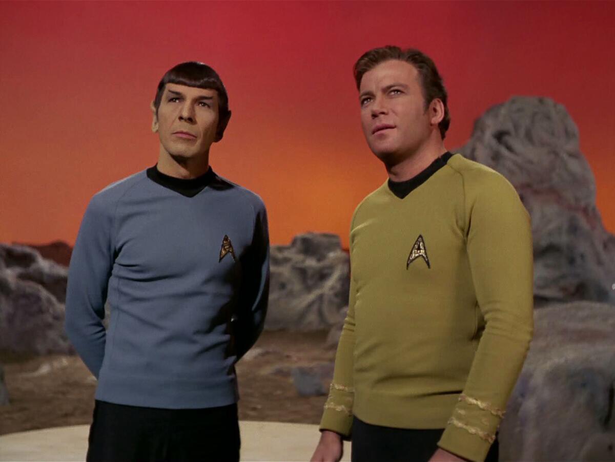 Leonard Nimoy in pointy ears and William Shatner stand on a TV set with craggy fake rocks and a red sky.