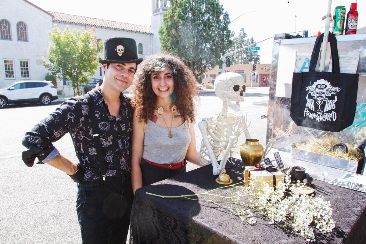A man and a woman in black clothing smile as they stand on a city street behind a hot dog cart and a plastic skeleton.