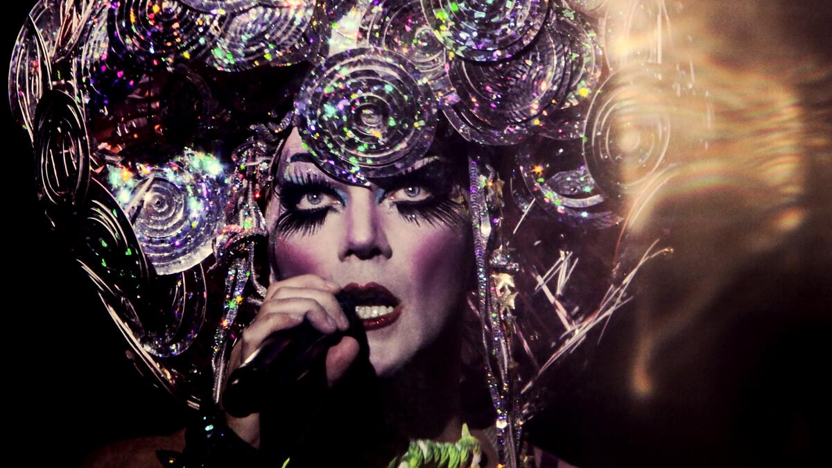 Taylor Mac in white and purple makeup and a silver glittering headdress, holding a microphone.