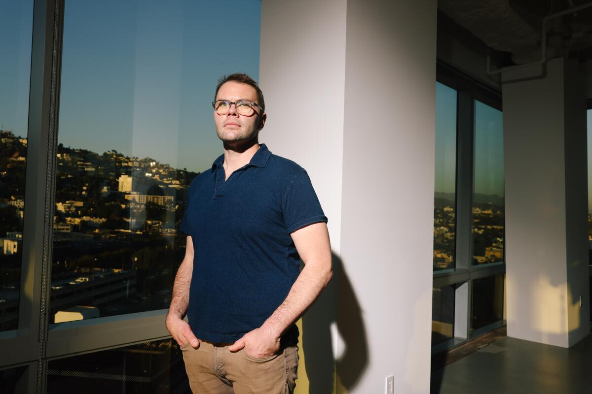 A man poses for a portrait in tan jeans and a navy shirt against a window looking over the Hollywood hills.