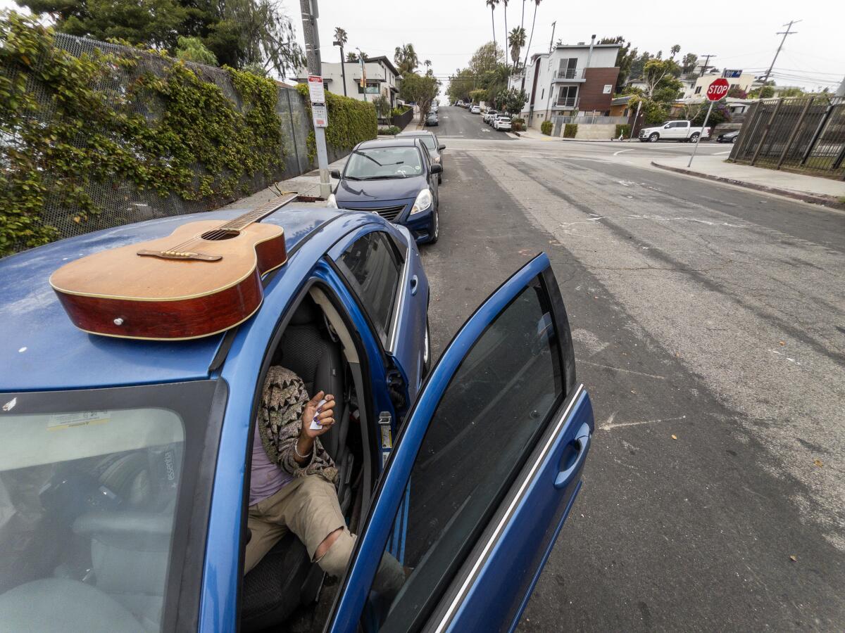 A woman, her face not visible, sits in the dirver's seat of a car, the door ajar as a guitar lies on the the car's roof.