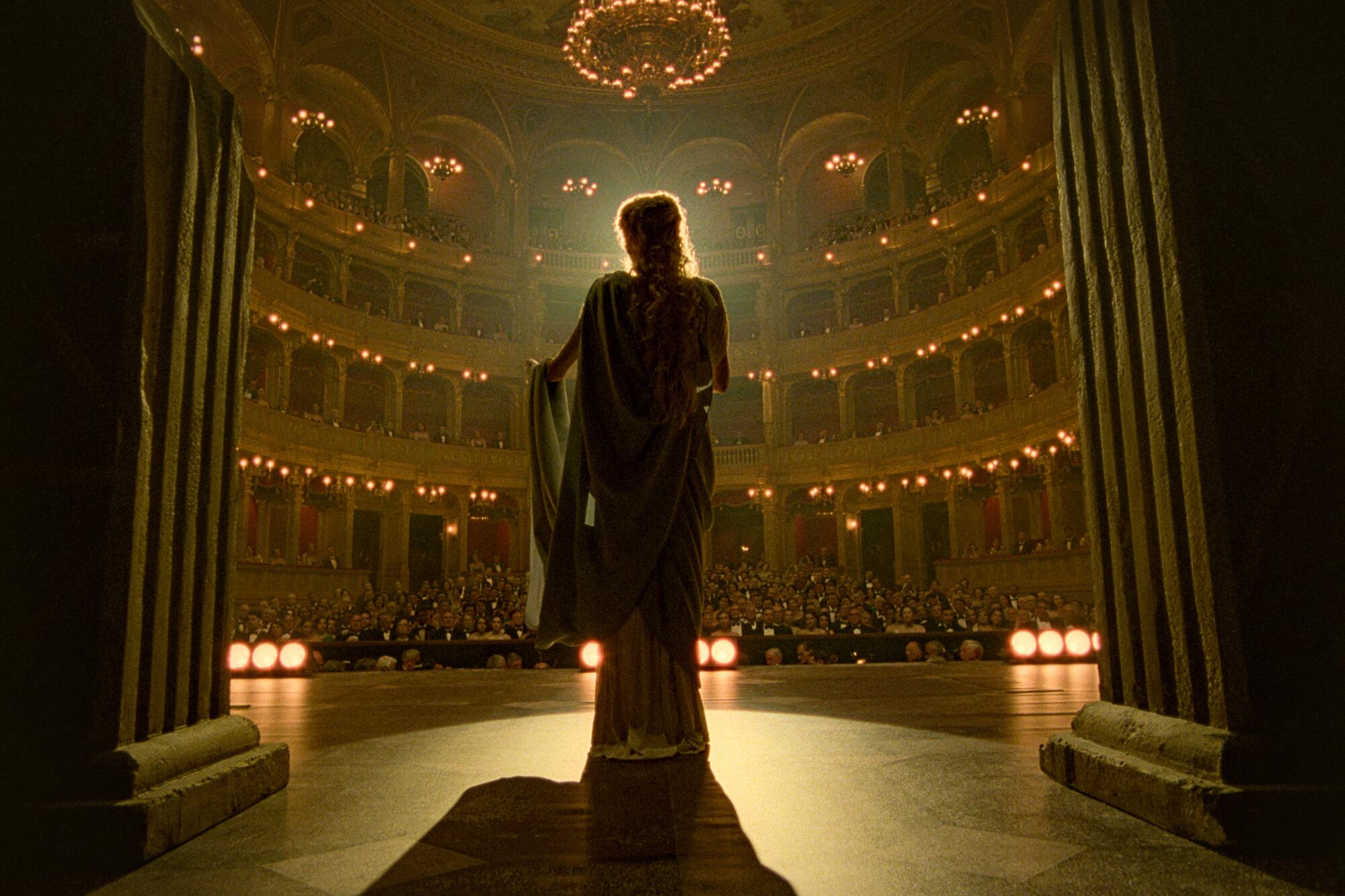 A woman in a gown is silhouetted from behind on the stage of a large theater in "Maria."