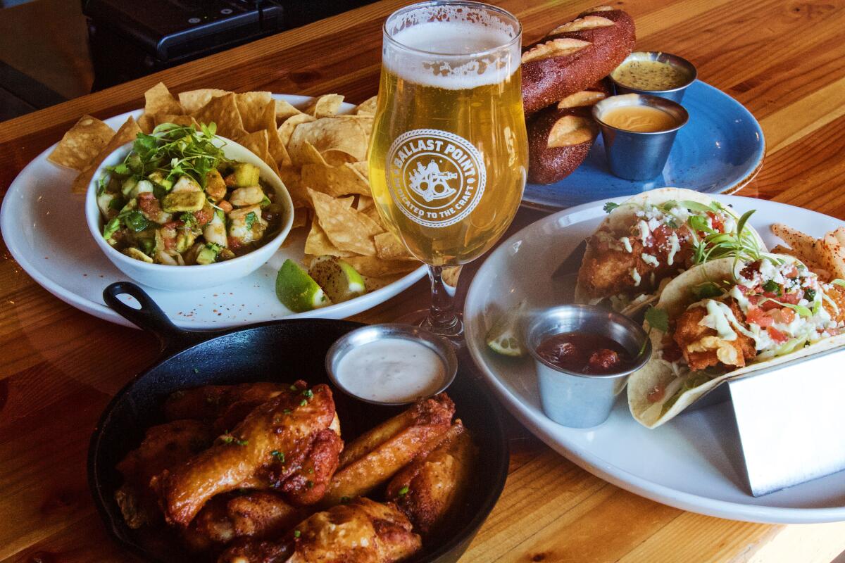 A tall golden beer in the center of a spread of food including aguachile with chips, skillet of wings and two fish tacos