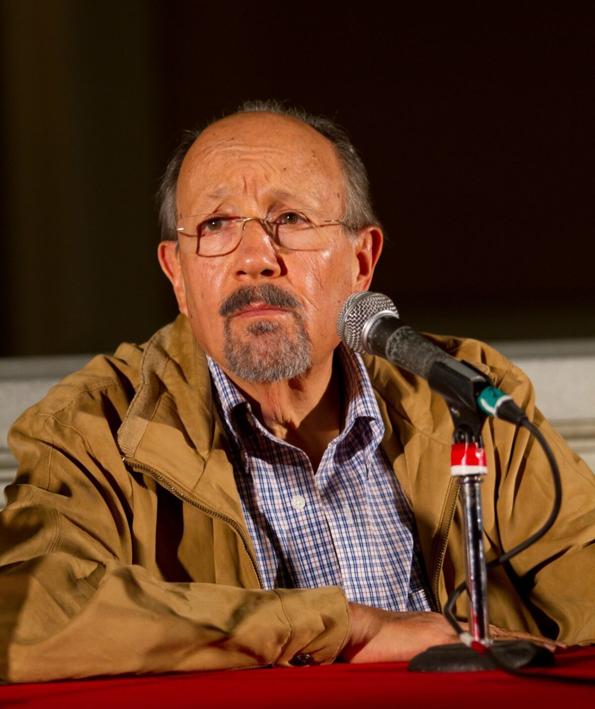 An elderly man in a brown jacket and a blue plaid shirt sits at a table with a microphone in front of him