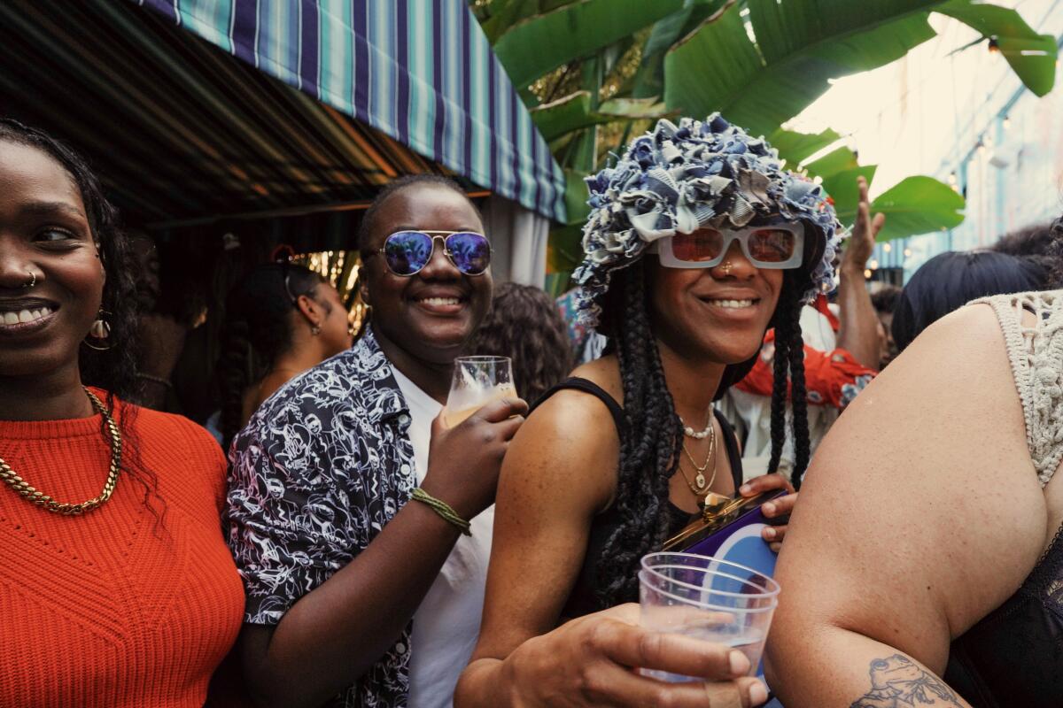 A group of Black people smile at the camera. In the center, a person wears chunky sunglasses and a ruffly hat. 