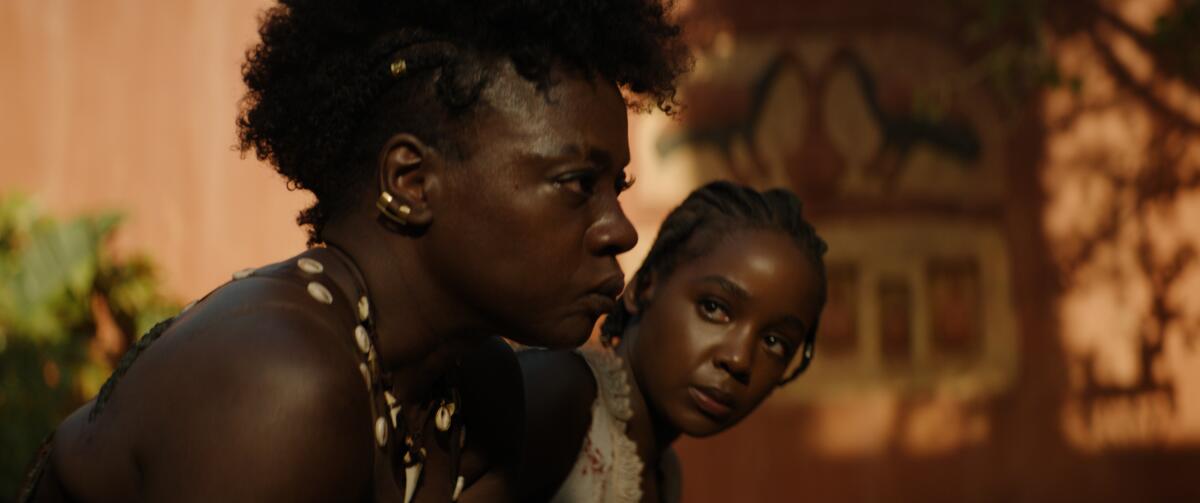 Two women sit side by side with somber expressions. One wears a necklace with shells and animal teeth.