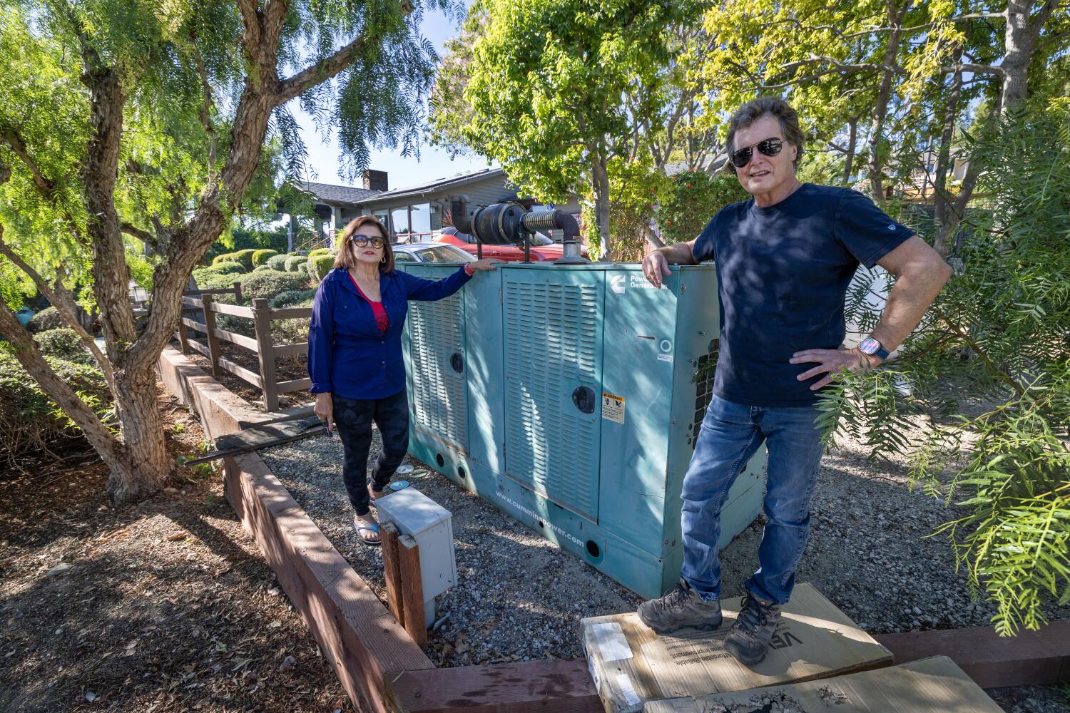 Rancho Palos Verdes, CA - October 30: Joe Badame and Martha de la Torre and her mom, Elvia de la Torre, not shown, who are still living without municipal electricity and natural gas service due to ongoing landslide movement in the area in Rancho Palos Verdes, pose with their propane generator. It's been more than a year, and Joe is worried about the winter and wants SCE to reenergize their neighborhood. His mother-in-law requires power during the night, which is hard on their solar panel. SCE says it's still too dangerous to reconnect power. Photo taken in Rancho Palos Verdes Thursday, Oct. 30, 2025. (Allen J. Schaben / Los Angeles Times)