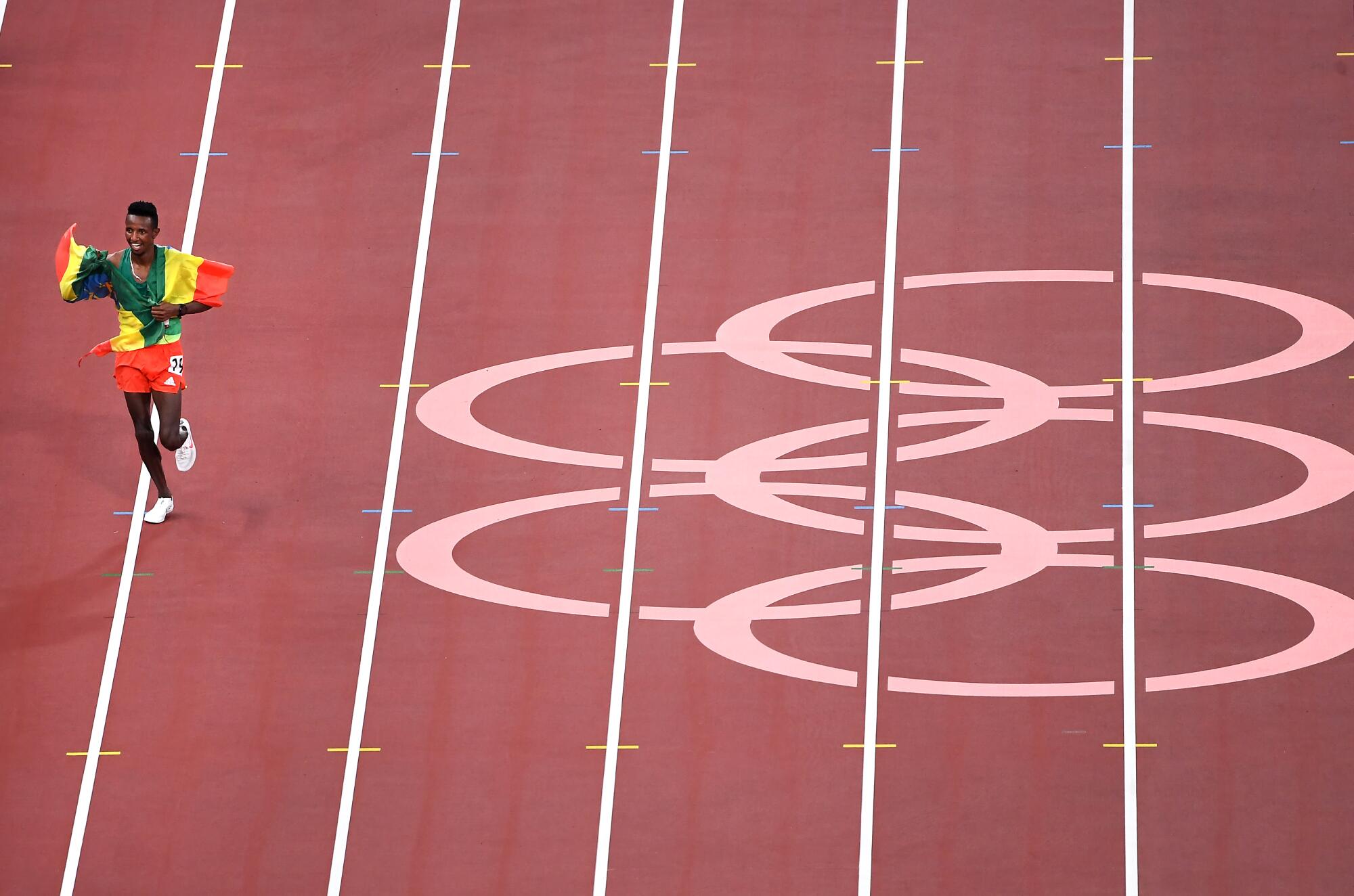 A man with a flag draped around his shoulders runs on the track past Olympic rings.