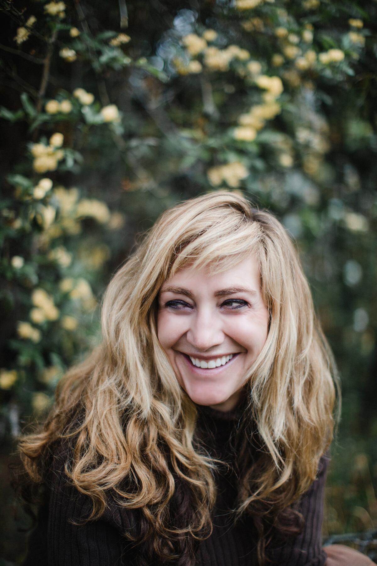 A blond woman with bangs smiles and looks to the left.