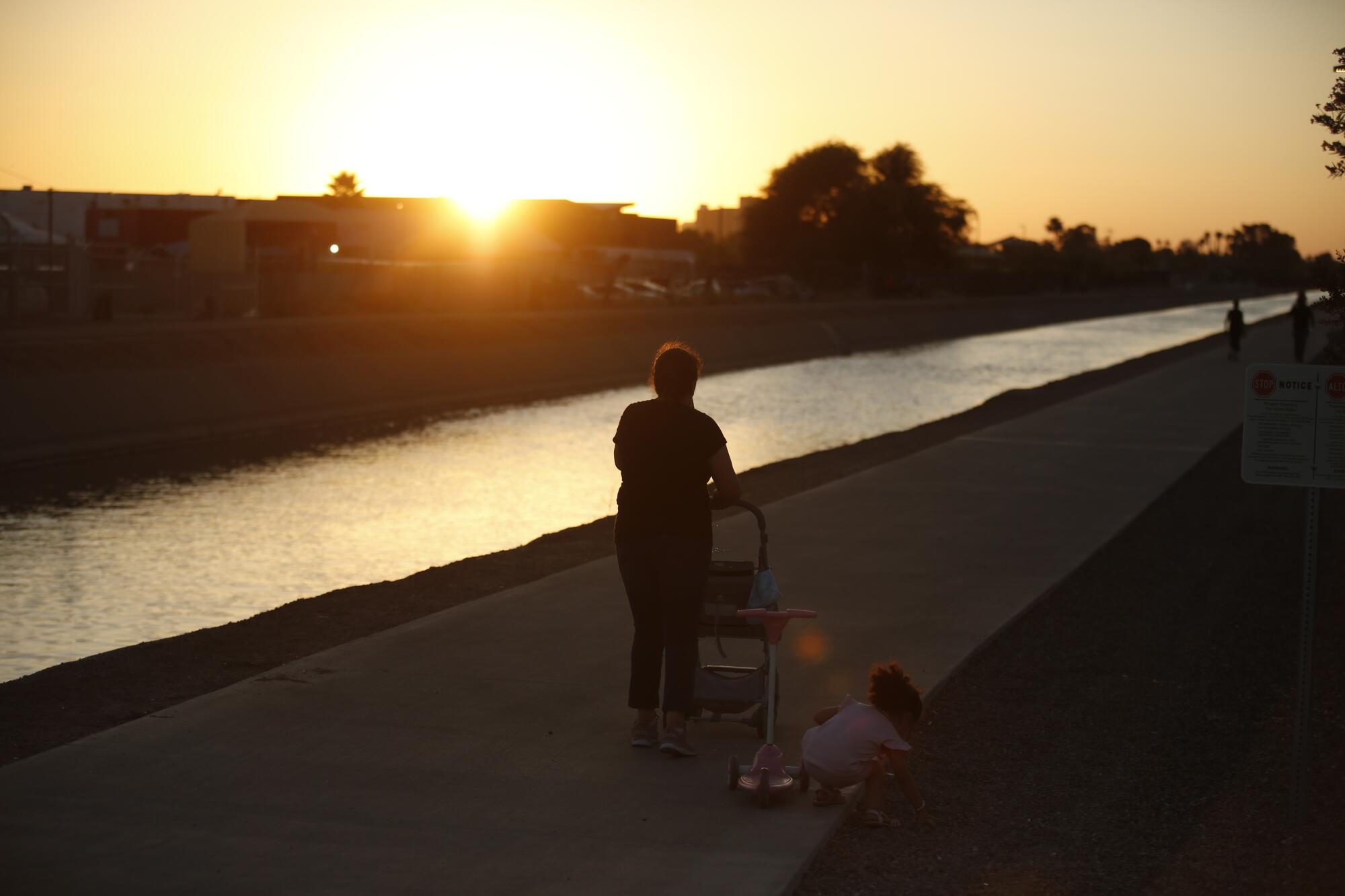 A toddler squatting and reaching for something off the path as she follows a woman walking along a waterway at sunset