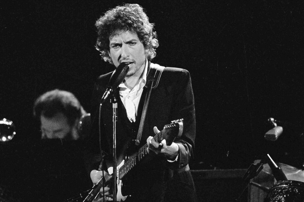 A black-and-white photo of Bob Dylan singing into a microphone and playing guitar onstage