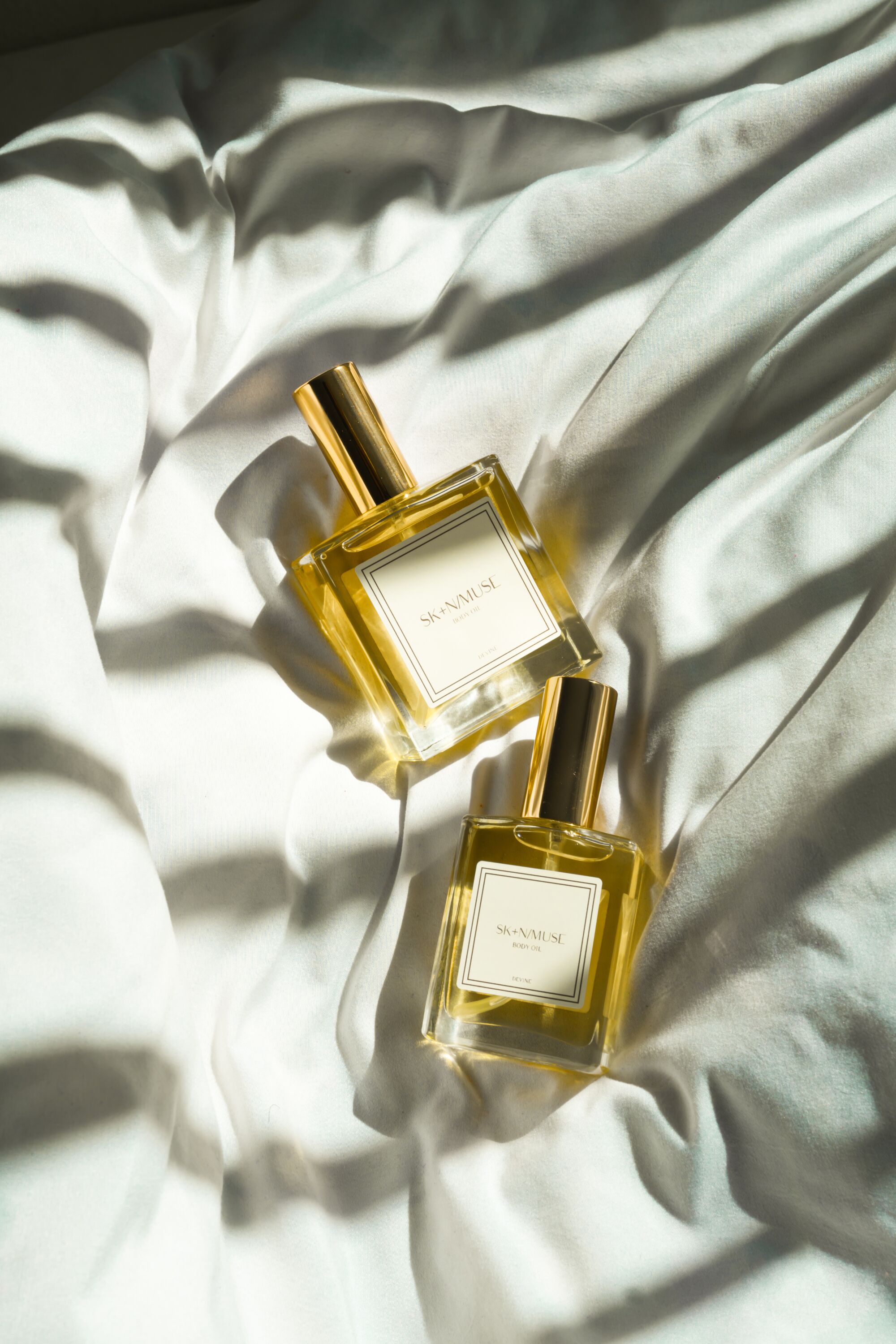 two SKNMUSE body oil bottles lying on white sheets with harsh window blind shadows cast on top of them