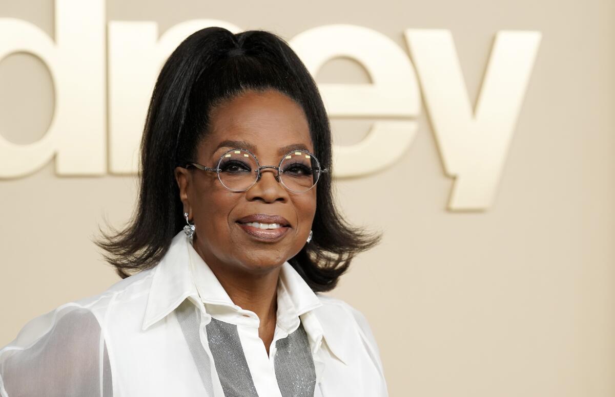 Oprah Winfrey smiles while wearing round glasses, a white blouse and her hair in a high ponytail.