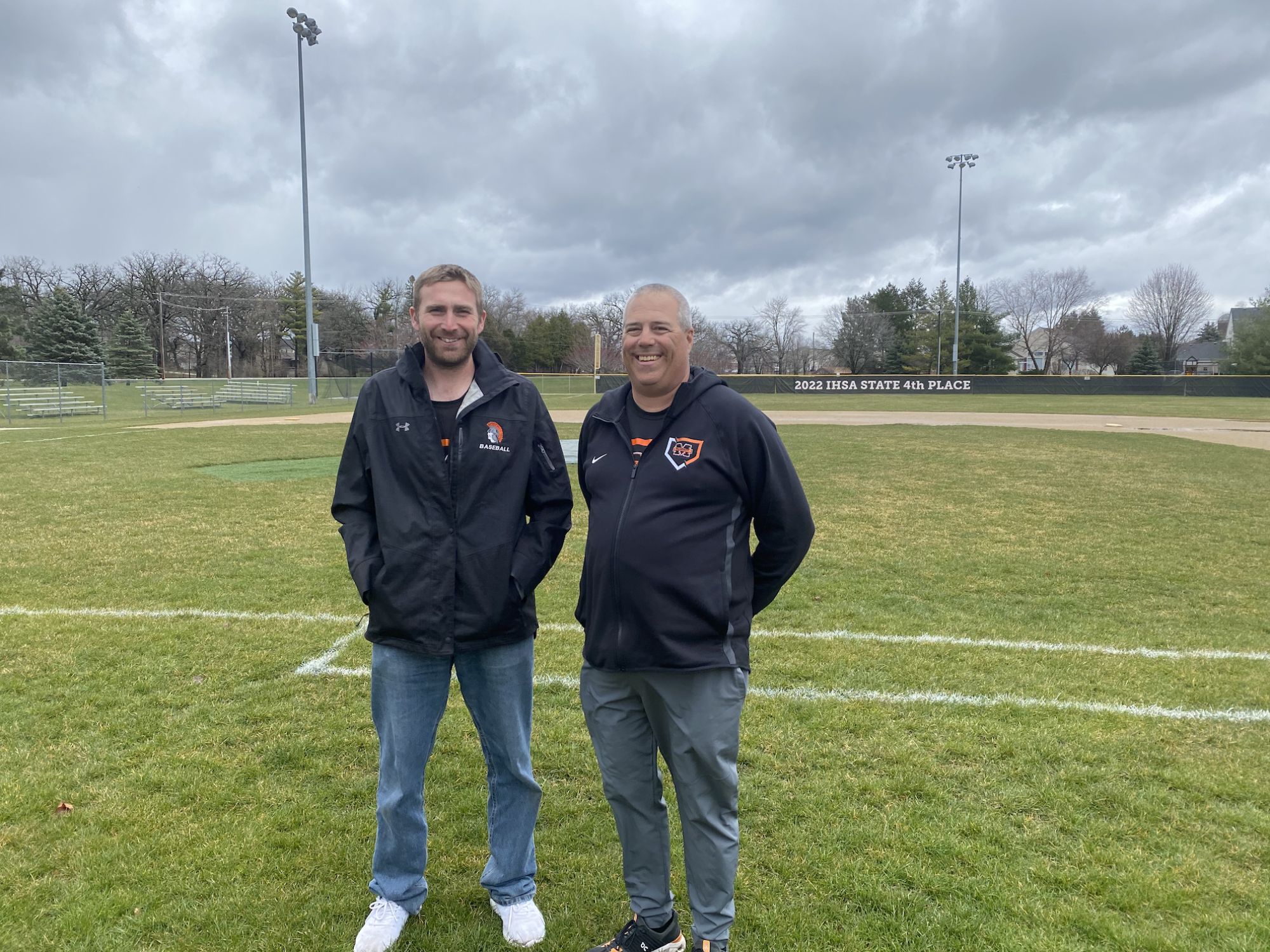 麦克亨利大学棒球队教练 Brian Rockweiler（右）和投球教练 Zach Badgley 在彼得森公园的球场上。