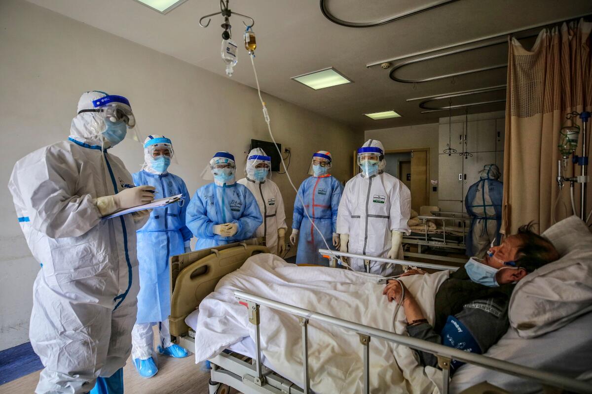 Medical workers in head-to-toe protective gear stand around a patient lying in a hospital bed