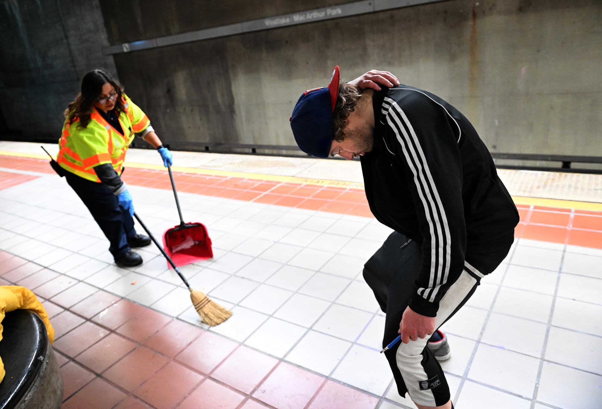 A man in a track suit, right, stands slumped while a woman wearing a neon vest sweeps a subway platform.