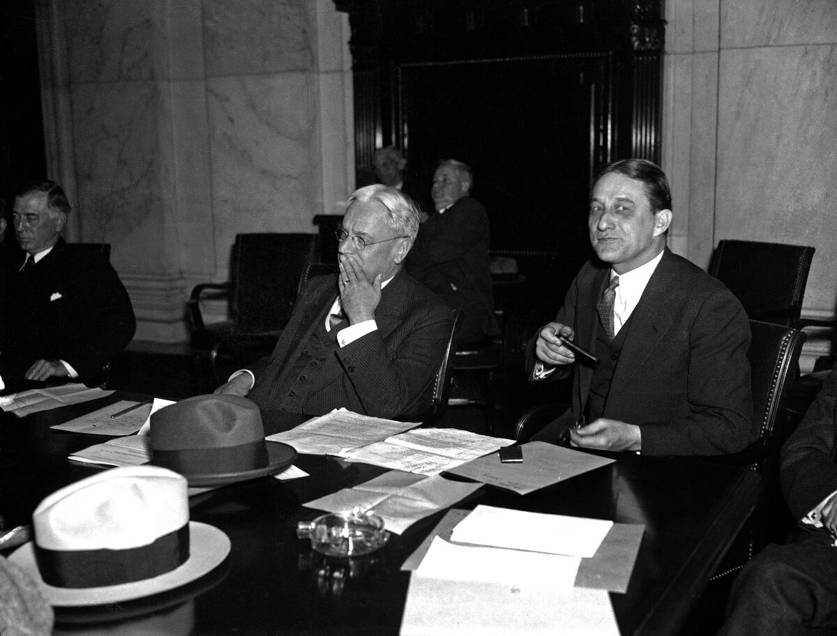 Hiram Johnson and Arthur Raymond Robinson, in three-piece suits, sit at a table that has papers, men's hats, an ashtray.