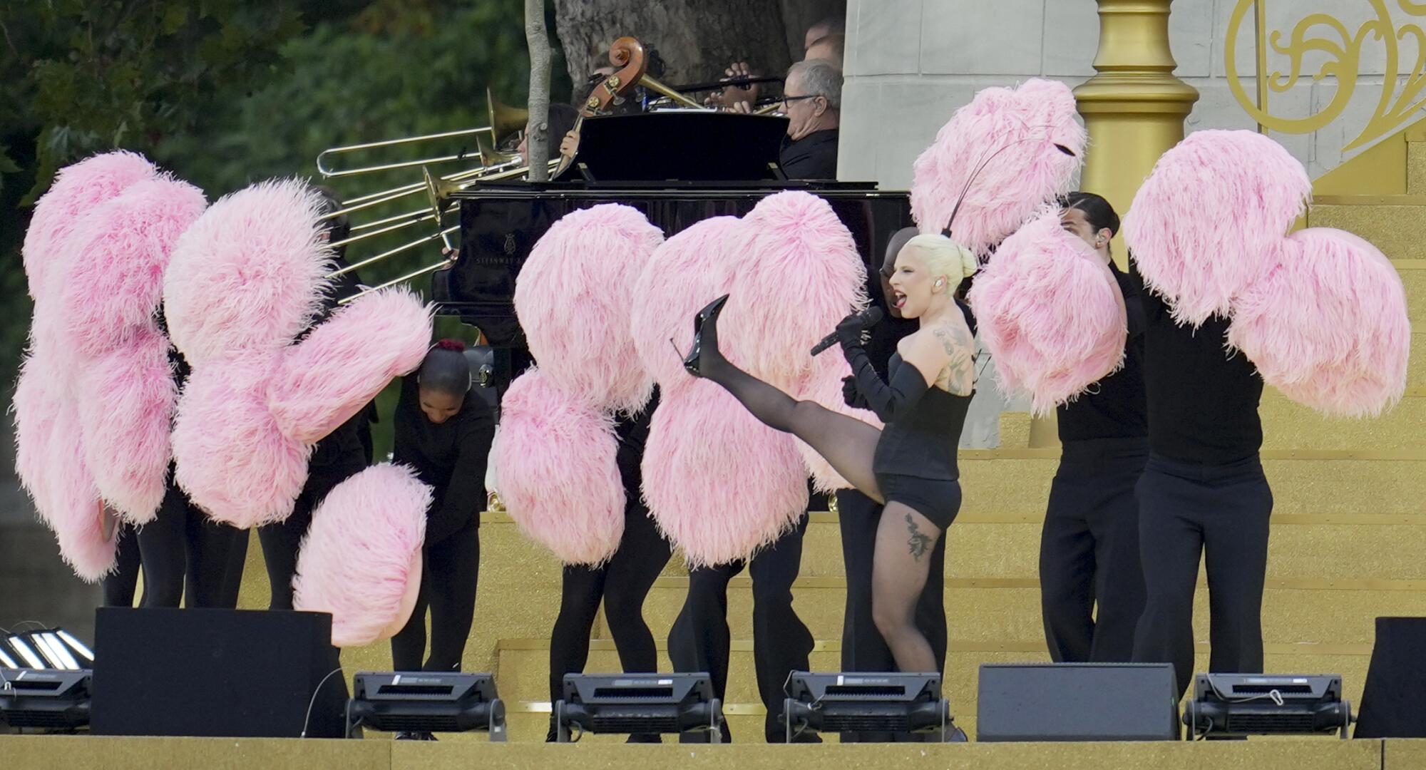 Lady Gaga kicking up a leg on a golden platform surrounded by dancers holding pink plumes.