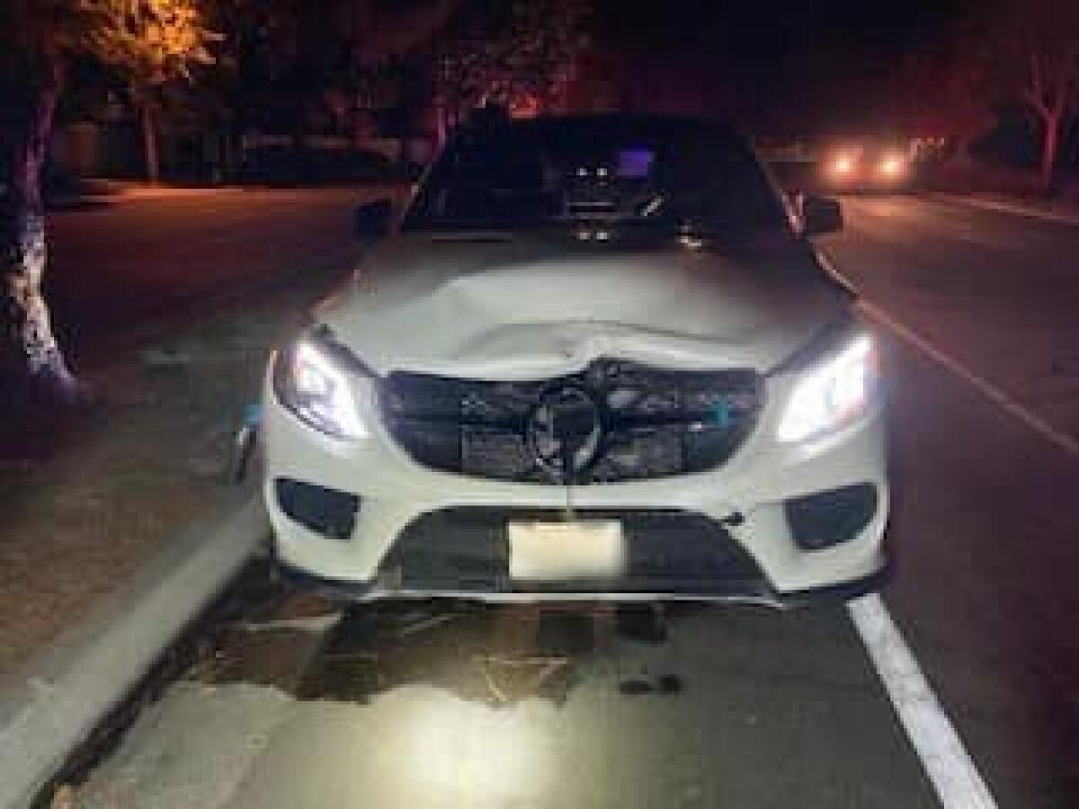 A white vehicle with a smashed hood and grille is parked alongside a road in the dark.